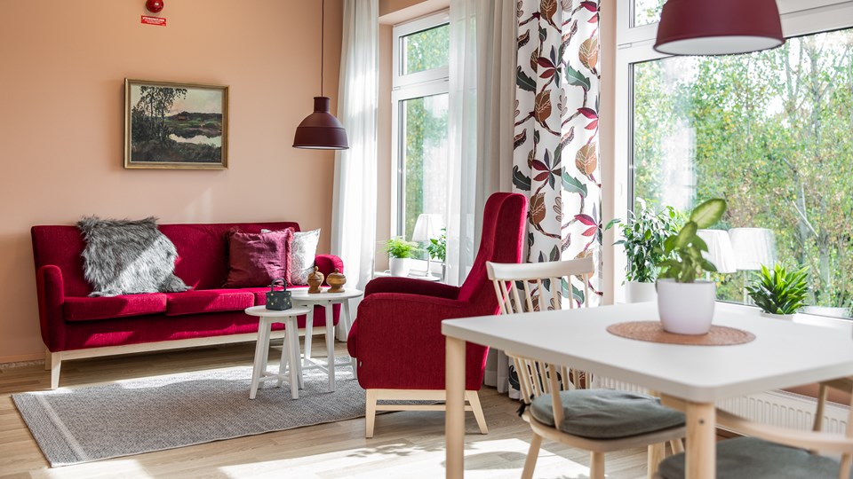 Ett ljust rum med stora fönster. Röd soffgrupp och ett matbord med stolar. Tavla, taklampor och mönstrade gardiner.