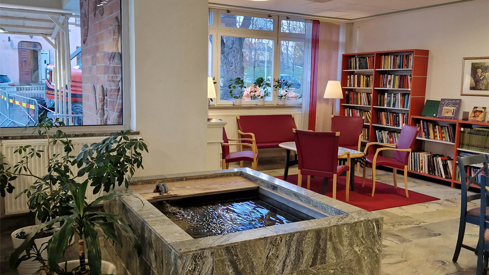 Stort rum med fönster mot gården. Röda bokhyllor med böcker finns utplacerade längs en vägg. Ett stort stenbelagt akvarium mitt i rummet.