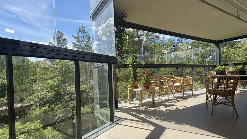Mindre sittgrupp med två rottingstolar och ett sidobord på en inglasad balkong. Utsikt över trädtoppar och närliggande byggnader under en klarblå himmel.