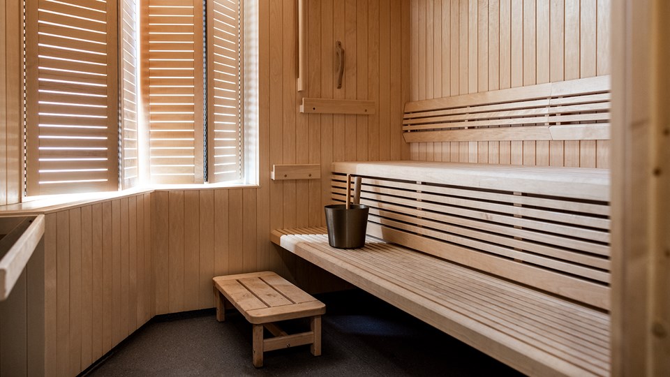 Bastu i trä med sittbänkar, fotbänk och ljus som sipprar från två fönster med träjalusier.
