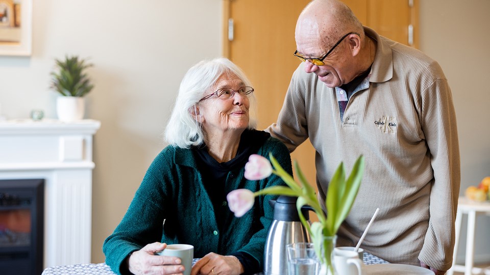 En man och en kvinna ler och samtalar vid ett bord dukat med kaffe och en vas med tulpaner.