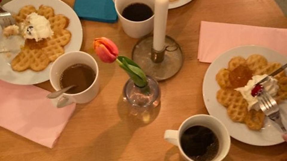 Tre personer sitter runt ett runt bord dukat med tallrikar och kaffekoppar. På tallrikarna ligger våfflor med sylt och grädde.