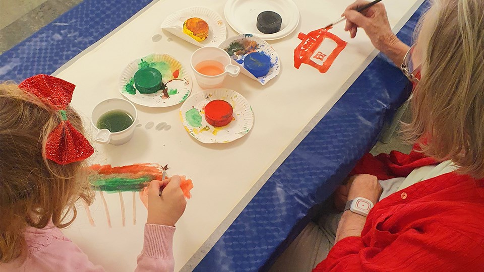 En äldre dam och en flicka målar tillsammans med vattenfärg. Ett långt papper är utrullat över bordet och målarfärger i olika färger finns utplacerade på bordet.