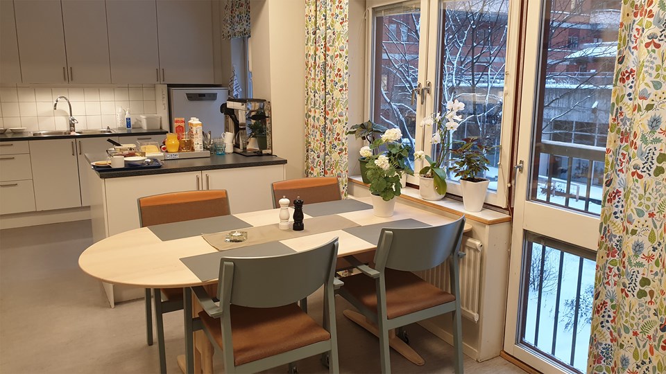 Ett ljust kök med sittplatser för fyra dukar med grå tabletter. Fönstren är prydda med växter. På köksön mellan kök och matplats står frukost framdukat.