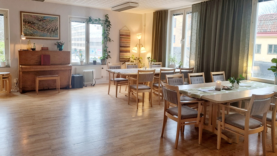 En ljus matsal med bord och stolar samt ett piano vid ena väggen. 