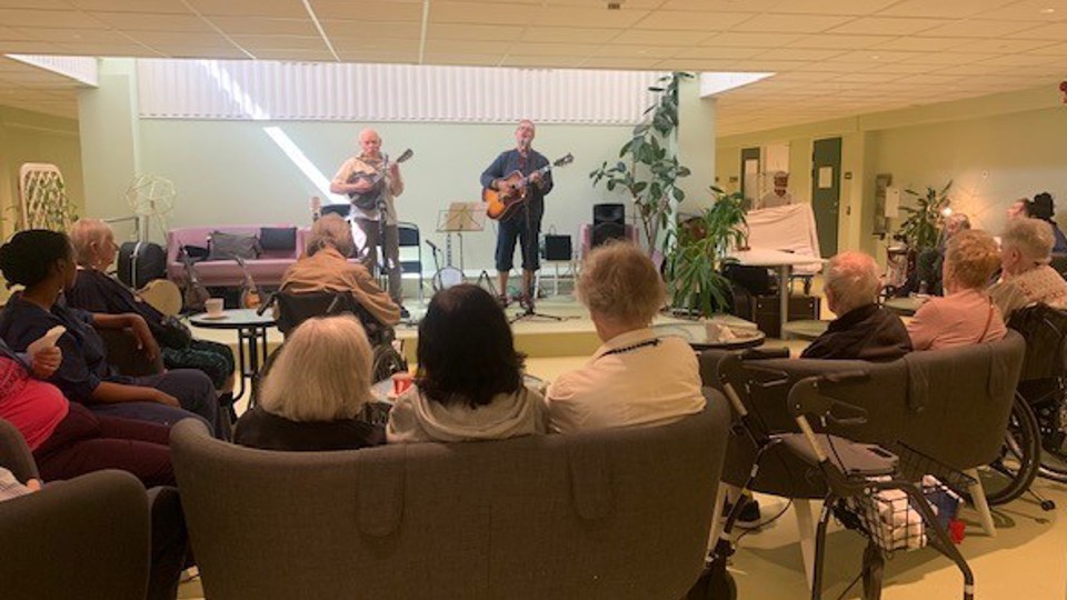 Seniorer sitter i Kista aktivitetscenter och lyssnar på två trubadurer