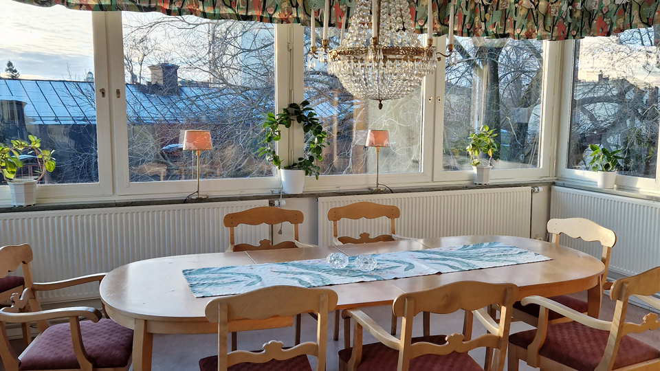 Bord med karmstolar står framför fönsterparti. I fönstren står några växter och två lampor med aprikosa skärmar.  I taket hänger en kristallkrona.