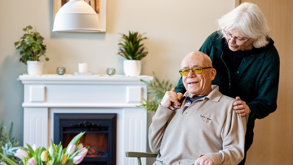 En äldre man sitter ned i ett rum med öppen spis och blommor i vas och krukor. En äldre kvinna står bakom mannen och håller honom i handen.