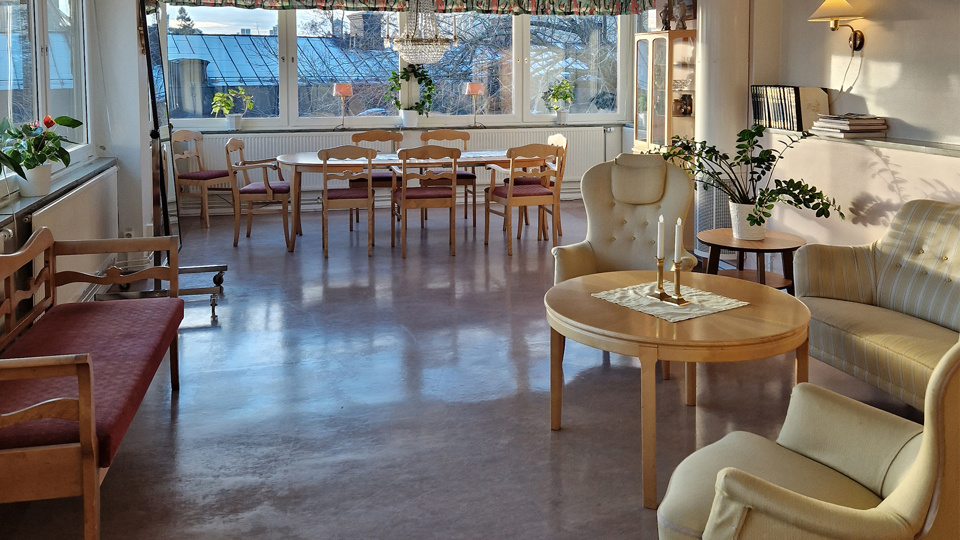 Salong med en beigegrön soffgrupp och ett träbord med två ljus på till höger. En pinnsoffa med en röd dyna till vänster och en matsalsgrupp längst bort vid fönstret. Fönsterparti sträcker sig längs två väggar.