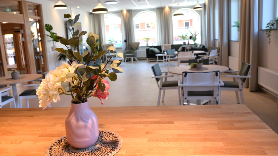 Ett rum med sittgrupper och stora fönster. I förgrunden en vas med blommor.
