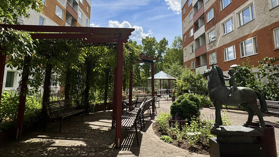 Innergård med pergolor, bänkar och grönska mellan två flerbostadshus. I förgrunden syns en skulptur av ett barn på en häst.