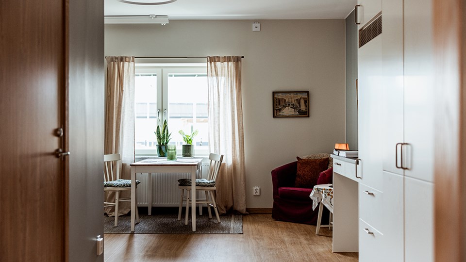 En hall med skåp och garderob och ett rum. I rummet syns en fåtölj, matbord med två stolar och ett fönster.