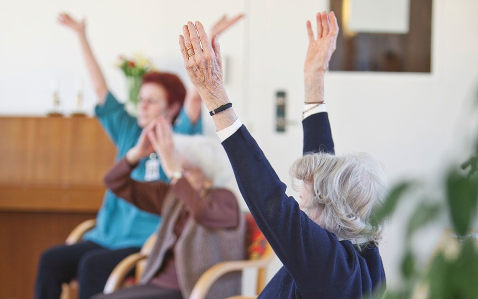Äldre kvinnor som gör sitt-gymnastik tillsammans och sträcker upp armarna.