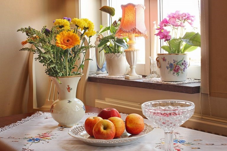 Bord med fruktfat, blomvas och skål med karameller. Fönster med krukväxter.