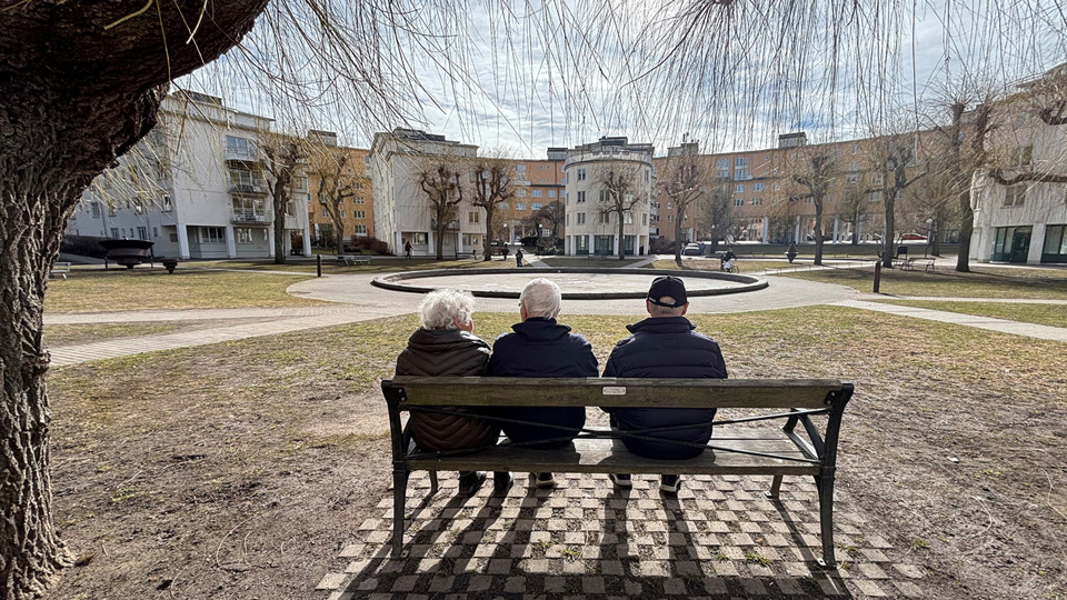 Tre personer sitter på en parkbänk med utsikt över en stor innergård omringad av hus.