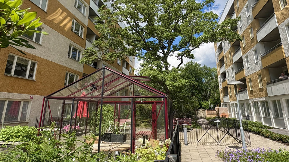 Inglasad gemensam terrass med sittgrupper i rotting och stora glaspartier från golv till tak. Terrassen omges av grönskande träd och släpper in mycket dagsljus.