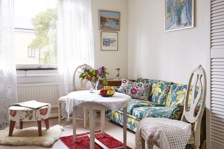 Ett rum med soffa, två stolar och en fotpall. Tavlor på väggarna och på bordet står blommor och ett fat med frukt.