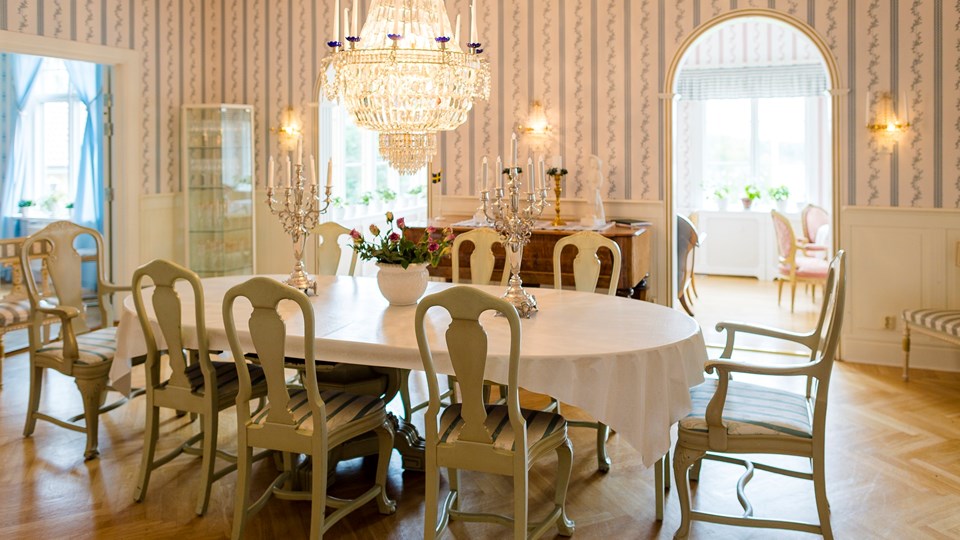 Matsal med möbler i herrgårdsstil, bord med vit duk, kandelabrar och kristallkrona.