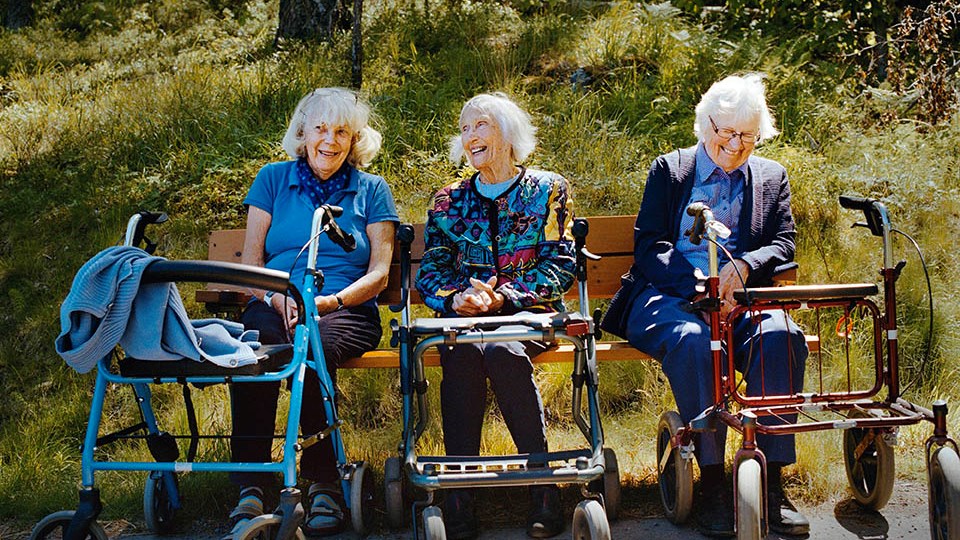 Tre seniorer sitter på en parkbänk och skrattar.
